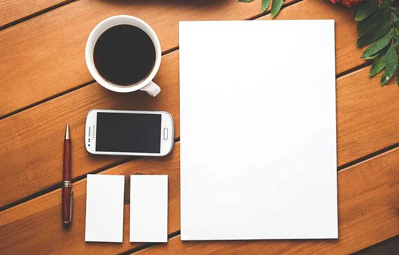 Wooden desk with a coffee, phone, pen, two blank business cards and a blank sheet of paper, ready for branding