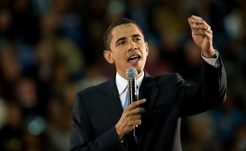 Man in a dark suit speaking animatedly into a microphone before a blurred crowd, like someone who dominates a conversation