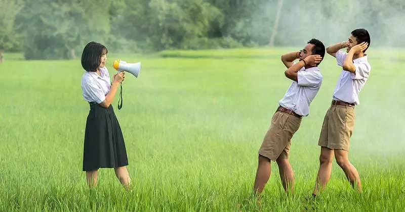 Girl shouting through a megaphone at two boys covering their ears in a green field, like a modern town crier