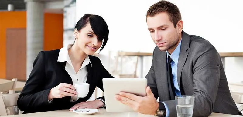 Two colleagues in business attire look at a tablet together over coffee, showing engaged teamwork in the workplace