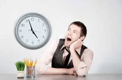 Bored young man in a vest and tie slumped at a desk, yawning as he watches the wall clock, showing a disengaged "whinger"